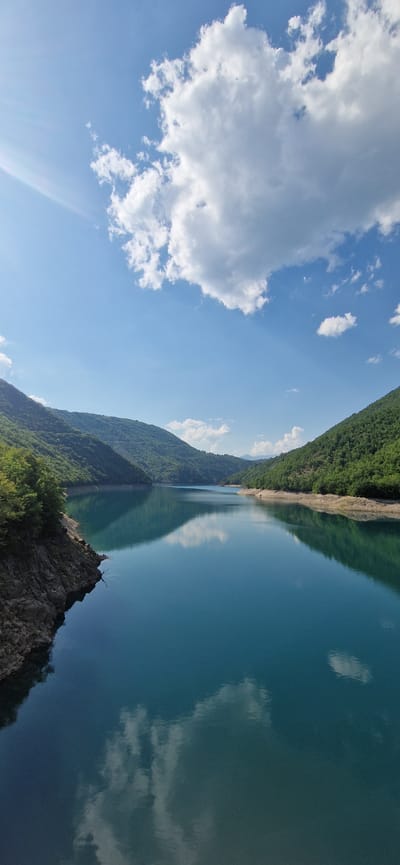The image captures a serene view of a tranquil lake in Bezuje, Montenegro. The water, a deep turquoise, is remarkably still, mirroring the dramatic blue sky above, dotted with fluffy white clouds. Lush green mountains flank the lake on either side, their slopes covered in dense vegetation. The left bank of the lake features a rugged, rocky terrain with sparse greenery, while the right bank is lined with trees. Sunlight streams from the upper left, casting a soft glow on the scene. The reflection of the clouds and mountains on the water's surface creates a sense of symmetry and depth. No people are visible in the frame. The environment appears undisturbed, conveying a sense of peaceful isolation.