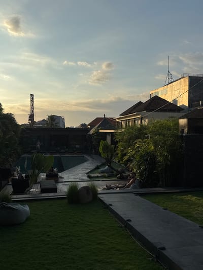The image shows an outdoor scene, seemingly in Pererenan, Indonesia, at sunset. In the background is a construction crane silhouetted against the sky, along with buildings of varying heights and architectural styles, some appearing to be hotels or resorts. In the midground is a swimming pool area with lounge chairs and greenery, including palm trees. A walkway, partially obscured by shadows, borders a grassy area. Two people are near the walkway, engaged in conversation or some interaction. The overall lighting suggests the warm tones of late afternoon.