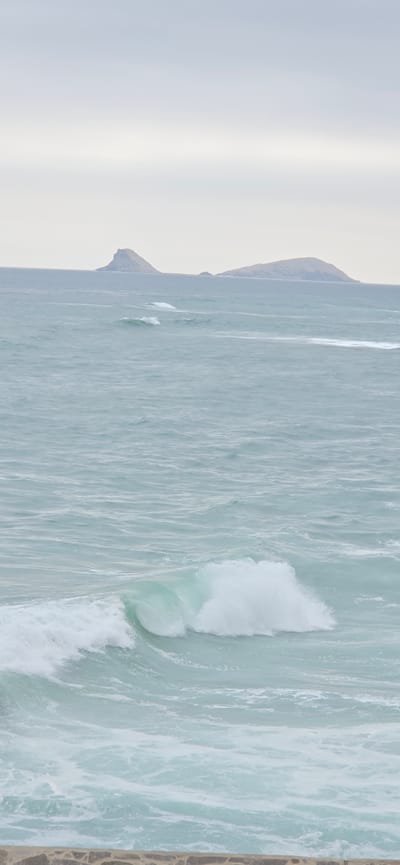 This image captures a serene seascape in Punta Hermosa, Peru. The foreground features a rocky embankment, possibly part of a seawall or natural coastline, constructed from irregularly shaped, light-colored stones. Beyond this embankment, the vast expanse of the ocean dominates the scene. Gentle, pale turquoise waves roll towards the shore, with white foam visible on their crests, indicating a moderate swell. In the middle ground, a larger wave is breaking, displaying vibrant white foam against the greenish water. The background reveals two distinct, muted islands or peninsulas on the horizon. These landmasses are low-lying and covered in a light brown or green hue, with their shapes softened by distance and atmospheric haze. The sky above is overcast, a uniform, pale gray that blends seamlessly with the horizon line, creating a tranquil and somewhat melancholic atmosphere. There are no people or animals visible in the frame, emphasizing the natural and undisturbed state of the environment. The overall impression is one of quiet coastal beauty, characteristic of Punta Hermosa's seaside landscape.