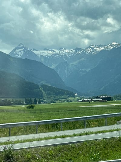 This is a landscape photograph taken in Zell am See, Austria, depicting a scenic view of mountains and a highway. The mountains, some with snow-capped peaks, dominate the left side of the frame. Their slopes are covered in dense green forests. In the foreground, a grassy field stretches out, bordered by a highway with two lanes and white markings. The highway runs parallel to the field, with a metal guardrail separating it from the grass. Scattered across the field are a few trees and some buildings, possibly farmhouses, with dark roofs. The sky is overcast with dramatic clouds, suggesting a cool or moody atmosphere. The time of day appears to be daytime, possibly late afternoon or early evening, given the lighting. The overall impression is one of natural beauty and tranquility, juxtaposed with the presence of modern infrastructure. There is no visible text in the image.