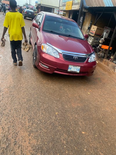 A person wearing a yellow t-shirt and black pants stands in front of a red Toyota car, holding two pieces of cloth. The scene appears to be a street in Barakin, Nigeria. The car has a license plate that reads "KWL-723GN ABUJA". Behind the car, there's a stall with various items, including boxes labeled "WP30X" and what looks like plumbing pipes. Signs for "SM Registration Center" and "LONDON PHONES" are visible in the background. The ground is wet, suggesting recent rain. The lighting indicates it's daytime, likely mid-morning or afternoon, given the presence of shadows. The overall atmosphere is that of a busy street or market area.