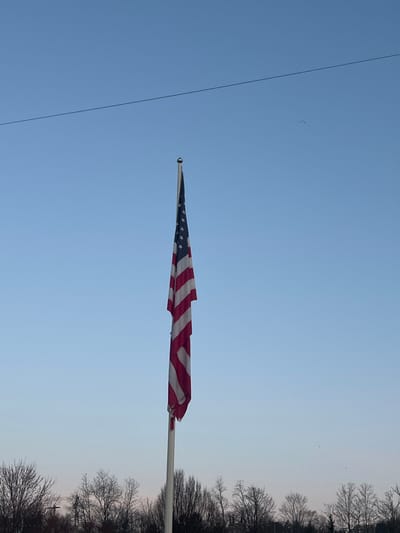 An American flag hangs vertically on a light-colored flagpole, situated in the center-right of the frame, beneath a clear, light blue sky in the Town of New Windsor, United States of America. The flag displays its red and white stripes and blue field with white stars, with minimal unfurling, suggesting calm atmospheric conditions. A thin, dark utility line crosses the upper portion of the sky horizontally. In the lower third of the image, a dense line of leafless trees forms the background, indicative of a colder season. No individuals are present. Several small, dark, indistinct objects are visible in the upper right quadrant of the sky.