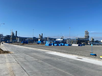 The image depicts an industrial construction site in the city of Ukhta, Russia, under a clear blue sky. In the foreground, a paved road made of large concrete slabs leads towards the background. The road has a textured surface and is bordered by gravel on the right and a dirt embankment on the left.

The middle ground of the image features a large industrial complex, characterized by a network of pipes, towers, and various structures painted in blue, yellow, and grey. Several blue modular buildings are also present, arranged in a line. Construction equipment, including a yellow excavator, is visible on the site. A few individuals in work attire can be seen in the distance, engaged in activities within the complex.

The background is dominated by the clear blue sky, with a few wisps of clouds present. The overall scene suggests an ongoing construction or expansion project at an industrial facility.