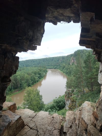 This media file shows a scenic view of a wide river flowing through a lush, forested valley. The perspective is from within a rocky overhang or cave, with rough, textured rock formations framing the scene. The river is a muddy brown color and curves to the left, bordered by dense green trees that cover the rolling hills on either side. The trees are a mix of deciduous and coniferous varieties, creating a rich tapestry of greens. In the distance, a rocky cliff face juts out from the treeline, adding a dramatic element to the landscape. The sky is overcast with a soft, diffused light, suggesting it is either early morning or late afternoon, or simply a cloudy day. There is no visible text, people, or objects in the frame, making the focus solely on the natural beauty of the location. The overall mood is one of tranquility and natural grandeur, inviting the viewer to imagine themselves in this serene wilderness. Based on the location context provided, this scene is likely in Al'kino, Russia.