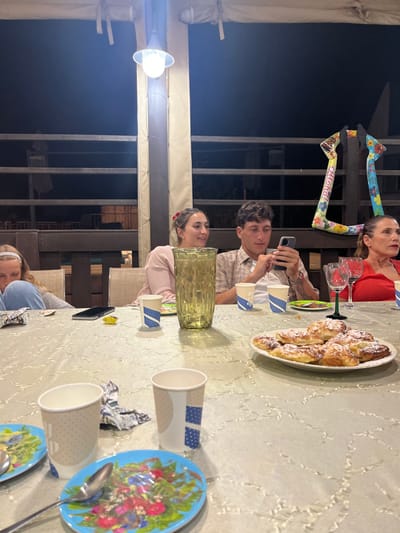 The image depicts an evening outdoor gathering on a patio or balcony in Beit Hanan, Israel. Four individuals are seated around a table covered with a light-colored, patterned tablecloth. From left to right: a young female, partially visible, wearing a light blue top and a white headband, looking downwards; a young female in a light pink top, smiling and looking to her left; a young male in a plaid shirt, intently viewing a smartphone held in both hands; and an older female in a red top, looking to her right.
On the table, a tall, ribbed, yellowish-green pitcher stands prominently. Multiple paper cups, some white with blue patterns and some plain beige, are present. Two blue plates with colorful floral patterns and spoons are visible in the foreground. A white oval plate piled with golden-brown pastries, dusted with powdered sugar, is positioned on the right side of the table. Two empty wine glasses, one with a clear stem and one with a green stem, are also on the table. Other items include a black smartphone, a small gold foil-wrapped object, and crumpled foil/paper.
The background shows a dark night sky, indicating evening. A wooden railing system defines the edge of the area, and light-colored fabric panels cover structural pillars. An overhead light fixture illuminates the scene. A colorful, balloon-like decoration spelling "Chana" (partially visible) hangs in the far right background. The general atmosphere suggests a casual social gathering or meal.