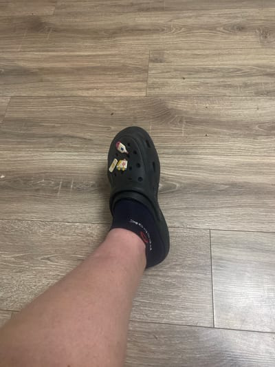An individual's left leg, wearing a black Croc shoe and a dark blue sock, is positioned on a wood-grain floor. The Croc shoe has two colorful Jibbitz charms attached: a thermometer and an ambulance with a band-aid. The sock has a red and white logo visible. The floor is composed of light brown planks with visible grain patterns and some minor scuffs. The scene is indoors, with no specific city or location information provided.
