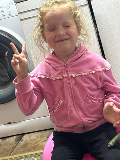 A young child with blonde curly hair is smiling with their eyes closed while holding up two fingers in a peace sign. They are wearing a pink zip-up hooded jacket with white ruffled trim. The child is seated in front of a washing machine and a light-colored cabinet, with a patterned rug visible on the floor. The scene appears to be indoors, possibly in a laundry area, in the city of Severnyy, Russia.
