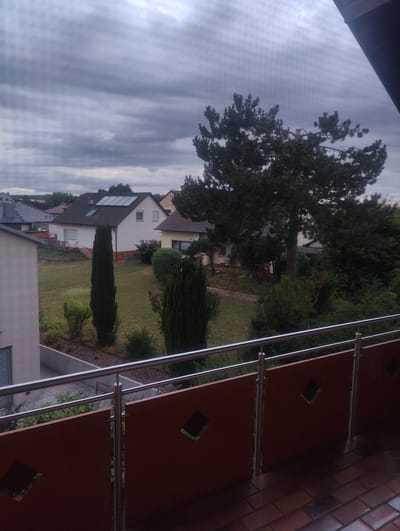 This is an outdoor, medium shot of a residential area under a cloudy sky, taken from a balcony. In the foreground, a brown balcony railing with diamond-shaped cutouts and a metal handrail is visible. Beyond the railing, a small lawn and garden area are present with several houses and trees. One house in the middle distance has solar panels on its roof. The sky is overcast with dark grey clouds.