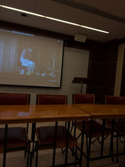 The image depicts an indoor venue, likely a classroom or conference room. The specific location within Tizapan, Mexico, is not discernible; however, text on the projection screen indicates "Nombre Sala : Salon211."
The environment consists of light-colored walls with dark wood paneling on the lower sections, a light-colored ceiling equipped with two linear fluorescent light fixtures and vents, and a tiled floor.
A large white projection screen on the left displays a scene from a video. The video shows a man in a light-colored collared shirt standing behind a counter, actively pouring liquid from a clear pitcher into a glass. A jug containing a yellow liquid and a white mug are visible on the counter. Above and to the right of the screen, a white wall-mounted speaker is affixed. Further right, a wooden lectern on a metal stand is positioned against the wall.
In the foreground, multiple glossy, light-brown wooden tables are arranged to form a larger surface. Surrounding these tables are chairs with dark red upholstered seats and backs, supported by black metal frames. No individuals are present within the physical room; only the figure within the projected video is depicted.