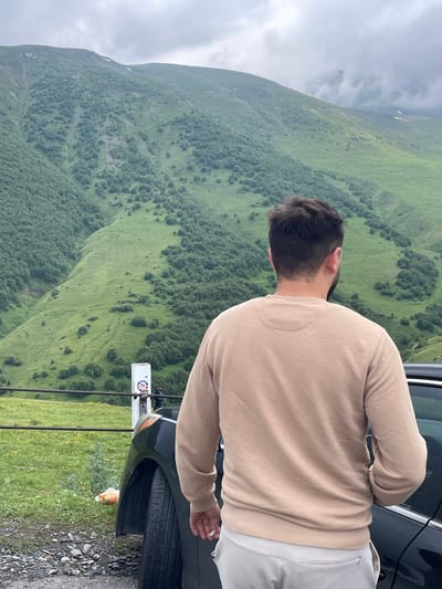 A man stands with his back to the camera, wearing a tan sweatshirt and light-colored pants, looking out at a lush green mountainside in Gergeti, Georgia. The mountain is covered in dense trees and scattered bushes, with a cloudy sky overhead. A black car is parked to the man's right, and a wire fence with a white post stands between him and the grassy slope. The post has stickers on it, and two people are visible in the distance on the other side of the fence. The ground in the foreground is a mix of gravel and grass, with a white plastic bottle visible near the car's front tire.