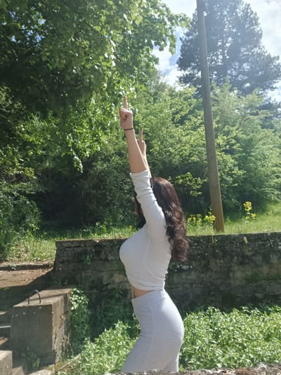 A woman is standing on a stone structure, potentially ruins, in what appears to be a natural, wooded area. She is wearing a light-colored long-sleeved shirt and gray leggings. She is facing towards the left of the frame with her arms extended straight out, pointing with her index fingers. The background consists of dense green trees and foliage, with a glimpse of sky and a wooden beam visible. The photograph may be taken in Gorna Verenitsa, Bulgaria based on context.