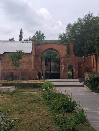 The image shows the entrance to a monastery in Martiros, Armenia, on a cloudy day. The entrance is an arched gateway made of red stone, with a black metal gate. To the left of the gateway is a building with a corrugated metal roof and red stone walls. A small tree stands in front of the building. To the right of the gateway is a tall, slender red stone structure topped with a cross. Beyond the gateway, lush green trees are visible. In the foreground, a paved walkway leads towards the entrance, bordered by grass and various plants, including rose bushes with red flowers.
