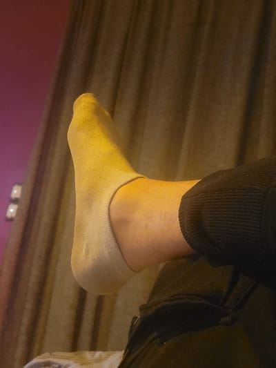 The image displays an elevated human left foot, clad in a light beige or off-white no-show sock. Visible hair is present on the ankle and lower leg. The individual is wearing dark, likely black, trousers with a ribbed texture, partially visible on the lower right. The scene is an indoor environment located in Ανδραβίδα, Greece. The background consists of a dark brown curtain with vertical pleats occupying the central and right portions of the frame. To the upper left, a magenta-colored wall is visible, featuring two small, indistinct light-colored points, possibly light fixtures or reflections. Artificial illumination casts a warm, yellowish glow primarily on the socked foot. No other individuals or discernible actions are present.