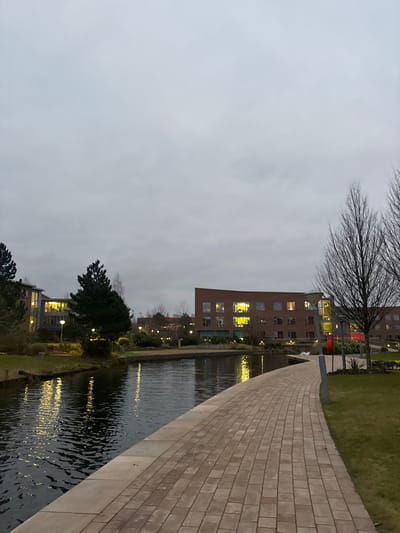 The image captures an outdoor scene at what appears to be dusk or early evening, characterized by an overcast sky and artificial lighting. The perspective is looking along a body of water, likely a canal or a narrow river, with buildings lining one side and a paved pathway on the other.

In the foreground, a wide, light-colored pathway made of rectangular paver blocks curves gently to the left, following the edge of the water. The blocks are arranged in a regular pattern. To the left of this pathway is the water, which reflects the artificial lights from the buildings and streetlights, creating shimmering streaks of yellow and white on its rippled surface. The water appears dark overall, consistent with the low light conditions.

Beyond the water, on the far bank, there are grassy areas interspersed with trees and shrubs. Several streetlights are visible along this bank, some of which are illuminated. Further back, a series of modern buildings with brick facades line the waterway. These buildings feature numerous windows, many of which are illuminated with warm yellow light, suggesting interior activity. One building, more prominent in the center, shows particularly bright yellow and some reddish lighting through its windows. The architecture appears contemporary, possibly residential or office buildings.

The sky above is uniformly light gray and overcast, indicating a lack of direct sunlight. The bare branches of a large tree extend into the top right portion of the sky, suggesting autumn or winter. Another cluster of dark green trees is visible closer to the buildings on the far bank.

No people or vehicles are visible in the scene. The overall impression is quiet and serene, with the artificial lights creating a warm contrast against the dim, gray surroundings. There is no visible text in the image.