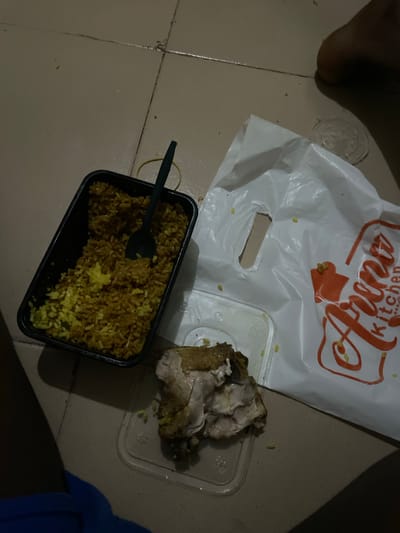 A overhead shot shows a plate of rice with chicken and a takeaway bag from "Amu Kitchen" on the floor. The floor appears to be tiled, with visible grout lines. A black plastic spoon is in the rice. To the right of the rice, there is a clear plastic container with a piece of cooked chicken. A white plastic bag with the "Amu Kitchen" logo is to the right of the food. The scene is dimly lit, casting shadows on the floor. The overall impression is of a casual meal setting on the floor, possibly in a residential area. It is not possible to definitively determine the location in Malete, Nigeria, from the image alone.