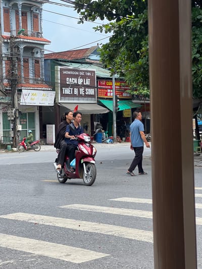The image captures a street scene in Tân Quang Commune, Vietnam, featuring a red motorcycle carrying two individuals in the foreground, a man walking on the right, and buildings lining the street. The motorcycle is a mid-shot, positioned on the right side of the road, with a man driving and a woman seated behind him, facing slightly to the left. The man is wearing a dark blue short-sleeved shirt and dark pants, while the woman is wearing a black outfit. A pedestrian, a man in a light blue short-sleeved shirt and black pants, walks away from the camera towards the right side of the image, crossing the street. In the background, several buildings are visible, including a multi-story structure with balconies and red-tiled roofs on the left, and other shops or businesses with signs. The street is paved and includes a pedestrian crossing marked with white stripes. The overall environment suggests a typical urban setting with ongoing daily activities.