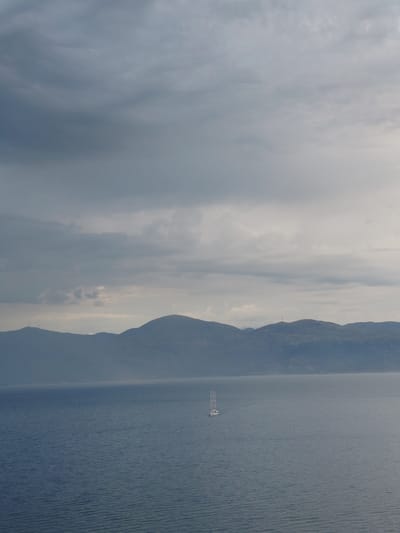 A sailboat with three masts is positioned in the center of a vast, rippling blue body of water, with a range of hazy, blue-green mountains forming a distant horizon line. The sky above is filled with a textured layer of grey clouds, some appearing darker and more dense than others, with patches of lighter grey and white sky peeking through. The overall atmosphere is muted and overcast, suggesting a cloudy or possibly stormy day. The scene is tranquil, with no visible people or other vessels. The location is the Municipal Unit of Aegio, Greece, specifically the waters facing its coastline.
