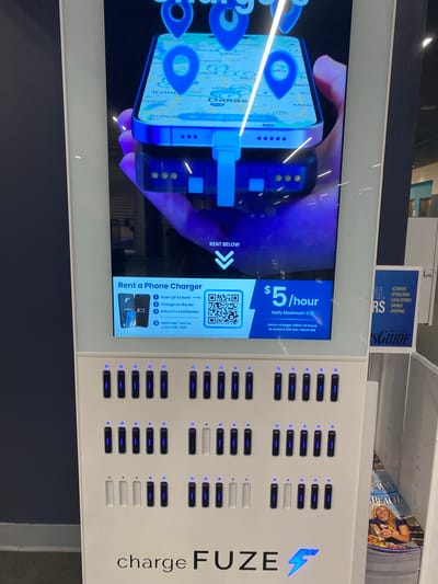 The image shows a ChargeFuze phone charging station located in Virginia Beach, United States of America. The charging station is white and features a digital screen displaying a map of Dallas, Texas with location pins. Below the screen, there is text explaining how to rent a phone charger, listing the cost as $5 per hour with a daily maximum of $30. There are multiple charging bays, most of which are occupied by dark-colored charging units with blue indicator lights. A few bays appear to be empty or occupied by white units. In the lower right corner, a magazine or brochure is visible with the word "BEACH" on it and a picture of a woman. The background is dark, suggesting an indoor environment, possibly a public space like an airport or mall.