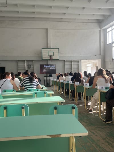 This media file captures an indoor scene, likely a gymnasium or large hall in Gladzor, Armenia. The focus is on a group of people, primarily women, seated at desks arranged in rows. They appear to be taking a test or participating in an exam, as indicated by the numbered labels on the desks, such as "27," "28," "29," "30," "31," "32," "33," "34," and "35." The individuals are mostly looking down, engrossed in their work.

The setting features light-colored walls and a wooden floor, characteristic of a sports facility. Natural light streams in from a large window or skylight at the top of the image, suggesting it might be daytime. A basketball hoop is visible on the wall in the background, further reinforcing the location as a gymnasium. On one of the walls, a large screen displays text that appears to be in a foreign language, possibly Armenian, given the location context. The overall mood seems focused and serious, typical of an examination environment. The desks are a light teal color with yellow metal frames. The people are dressed in casual attire.