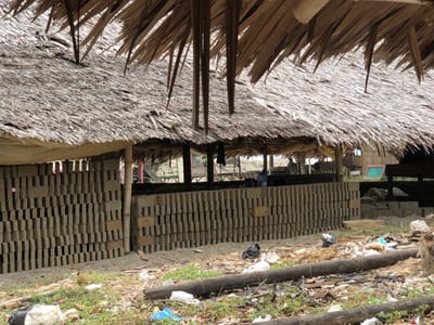 The image depicts an outdoor scene in Bangkajaya, Indonesia, likely a brick-making or storage area. The dominant feature is a thatched roof, constructed from dried palm fronds, providing shade over a sheltered area. Beneath the roof, tall stacks of grey bricks are neatly arranged, forming walls. The bricks appear to be of uniform size and shape, suggesting they are manufactured goods.
The ground in front of the brick stacks is covered in sand, scattered debris, and sparse green vegetation. Several pieces of trash, including plastic bags, are visible on the ground, indicating a somewhat unkempt environment. A fallen log lies horizontally across the foreground, further adding to the rustic and possibly industrial nature of the location.
In the background, through the openings of the sheltered area, a sandy beach or open area is visible, with further wooden structures and what appears to be a water cooler. The lighting suggests it is daytime, possibly overcast or late afternoon, as there are no harsh shadows. The overall impression is of a functional, utilitarian space in a tropical or subtropical climate. There are no identifiable people actively engaged in work within the frame, though one person is partially visible in the far background to the left. The scene conveys a sense of industry and everyday life in a rural or semi-rural Indonesian setting.