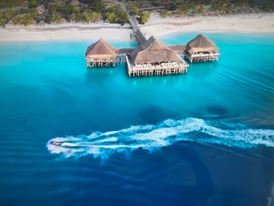 The image shows an aerial view of a tropical scene. In the foreground, a personal watercraft is creating a large white wake on dark blue water. Above the watercraft, there is an area of lighter blue water with a complex of buildings raised on stilts. The structures are connected to the shore by a long pier. The shore is lined with white sand and palm trees. In the background, there are buildings. The image does not depict any recognizable location within the city of Basingstoke and Deane, United Kingdom.