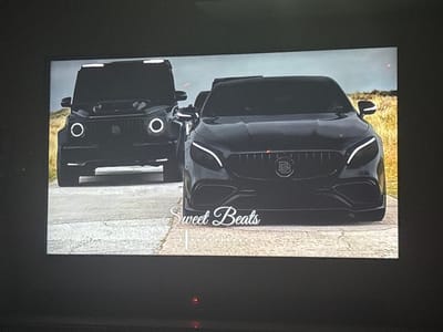 The image displays a photograph of a screen, which features two black Brabus-tuned Mercedes-Benz vehicles. A Brabus G-Wagen SUV is positioned in the background on the left, with its circular daytime running lights illuminated. A Brabus coupe, likely an S-Class variant, is in the foreground on the right, showcasing its distinctive multi-line LED headlights. Both vehicles are stationary, facing forward, and appear to be parked on a light-colored paved or dirt road. The background consists of dry, light-colored vegetation, consistent with a field or grassy area, under a pale sky. White text reading "Sweet Beats" is overlaid on the lower central portion of the image. No individuals or specific actions are depicted. The visual information within the image does not contain identifiable landmarks or details to confirm the location as Borisovo, Bulgaria.