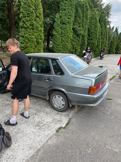 In Stara Rafalivka, Ukraine, a young man in a black t-shirt and shorts stands next to the open driver's side door of a gray sedan. He is looking away from the camera, towards the right side of the frame. The car, a Lada sedan, is parked on an asphalt surface next to a row of tall, dense green cypress trees. In the background, a person is seated on a dirt bike, and another person is visible on a scooter further down the row of trees. The ground is a mix of asphalt and a lighter colored aggregate material with scattered patches of grass. The overall scene suggests an outdoor gathering or casual stop, possibly in a park or recreational area.
