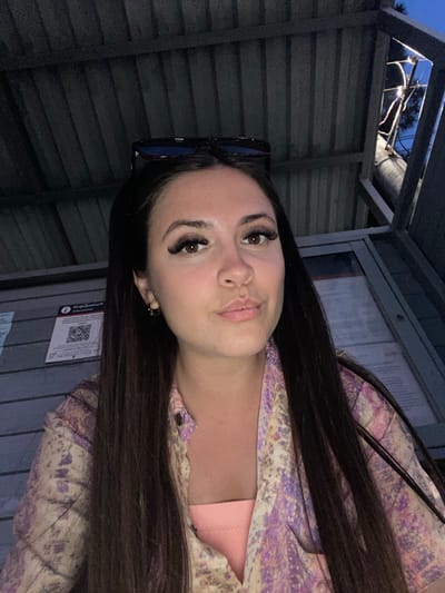 A young woman with long, dark hair is pictured outdoors at what appears to be a bus stop shelter. She is wearing a light pink top and a patterned shirt with purple and yellow floral designs. Sunglasses are perched on top of her head. In the background, there is a grey corrugated wall, a display case with information, and a tree with some wiring visible against the twilight sky. A QR code is visible on a notice posted on the wall. The image is lit by artificial light sources, contributing to the overall low-light atmosphere.