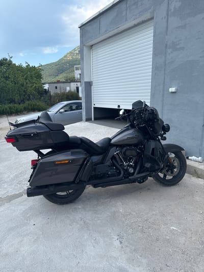 A dark grey Harley-Davidson motorcycle is parked on a concrete surface in front of a grey building with a large, white, ribbed garage door. The building is located in Radanovići, Montenegro. To the left of the motorcycle, a silver sedan is parked behind it. In the background, there are green trees and a hilly, mountainous landscape with scattered buildings. The sky is cloudy with patches of blue. A white bottle is lying on its side on the ground to the right of the motorcycle.
