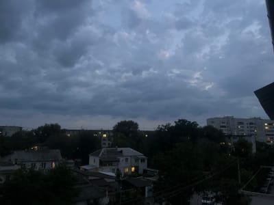 This is a night scene in Saky, Ukraine, with a cloudy, overcast sky. The foreground shows the roofs of several buildings, some with visible windows and lights on. In the mid-ground, there are trees and houses, with some windows illuminated. In the background, a multi-story apartment building is visible with lights on in many of its windows. Power lines are also present in the scene, along with a row of parked cars on the right side. The overall impression is a residential area at dusk or night, under a stormy sky.