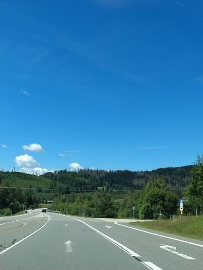 The image depicts a scenic road in Strážný, Czechia, on a clear, sunny day. The road curves to the left and right, with white lane markings guiding traffic. A dark-colored car is visible on the left lane of the road in the distance. The roadside is lined with lush green trees and rolling hills covered in dense forest. The sky is a vibrant blue with a few scattered white clouds. Several road signs are present, including a blue sign with a white arrow pointing upwards and a red circle with a horizontal line, indicating a prohibition. Another triangular sign with a red border is also visible. The overall impression is of a tranquil, natural landscape.