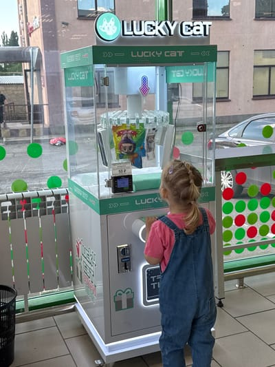 The image shows a young girl in a pink shirt and denim overalls playing with a "Lucky Cat" claw machine. The machine is white and green with colorful circles on the glass. There are prizes inside the machine, including a bag of "Labubu" toys. The girl is looking intently at the machine, her hands near the controls. The machine has text in Russian, including "СИМУЛЯТОР ЛОРКОСТИ" ("Simulator of luck") and "50 P" (likely the price per play). The setting appears to be outdoors, possibly in a shopping area or entertainment venue, given the reflection of buildings and trees in the glass of the machine. The lighting suggests it is daytime. The girl's focus and the presence of a game suggest she is engaged in playing and enjoying herself. The machine itself is a prominent object, decorated with the "Lucky Cat" logo and branding.