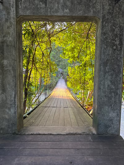 The image shows a wooden suspension bridge seen from within a concrete archway. The bridge spans across a body of water, with lush green foliage on either side. The sunlight filters through the leaves, casting a warm glow on the wooden planks of the bridge, which appear weathered and worn. The bridge extends into the distance, where it leads to a small structure with a thatched roof, possibly a pavilion or a small building. The concrete archway frames the view of the bridge and its surroundings, creating a sense of depth and perspective. The overall scene suggests a tranquil, natural setting, likely a park or a nature reserve. The presence of the suspension bridge and the surrounding greenery could be characteristic of natural attractions in Ranong, Thailand, known for its tropical landscapes and rainforests. There are no people visible in the image. The scene is static and peaceful.
