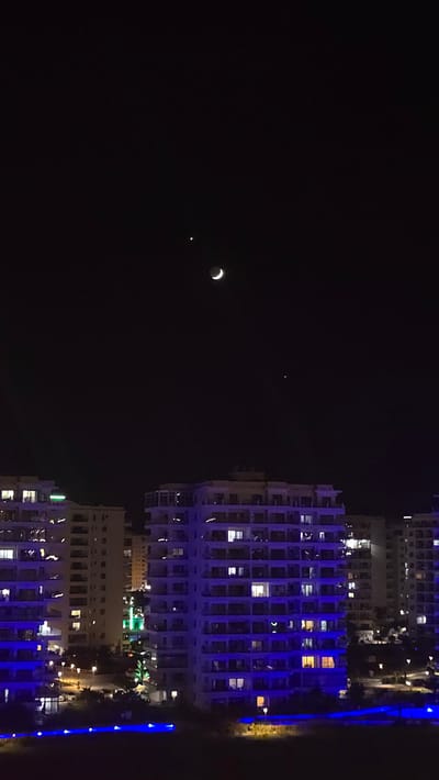The image depicts a nighttime scene of a crescent moon and a small, bright star or planet in a dark sky, with several tall buildings illuminated by artificial lights in the foreground. The buildings are multi-story structures with numerous windows, many of which are lit, suggesting occupancy or interior lighting. Blue and yellow lights are visible along the exteriors of the buildings and in the surrounding area, creating a vibrant, urban nighttime atmosphere. The orientation of the image is rotated, with the buildings appearing to rise from the bottom and extend upwards towards the sky. The sky is predominantly black, with the celestial bodies standing out against the darkness. The presence of the moon and a bright celestial object suggests it is nighttime, likely clear weather for visibility. The illuminated buildings and their architectural style hint at a resort or residential area, possibly along a coastline, given the dark expanse to the right which could be water or land. No people are distinctly visible, but the lit windows imply human activity. There is no visible text in the image.