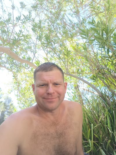 The image shows a man from the chest up, outdoors, with a background of green foliage and a bright blue sky. He has short brown hair and blue eyes and is smiling. His chest and shoulders are bare. The foliage appears to be from trees and tall grasses. The lighting suggests it is daytime. The man is positioned in the foreground, with the natural environment filling the rest of the frame. The location is the city of Ontinyent, Spain.
