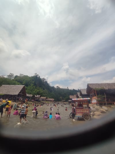 A group of people are enjoying a day at the river in Pantonrayeuk Satu, Indonesia. The river is shallow and many people are wading in the water. There are several huts with thatched roofs on the riverbank, and a food cart selling crispy buloh. The sky is cloudy, but the sun is shining through in some areas. The river is surrounded by lush green trees and a hilly landscape.