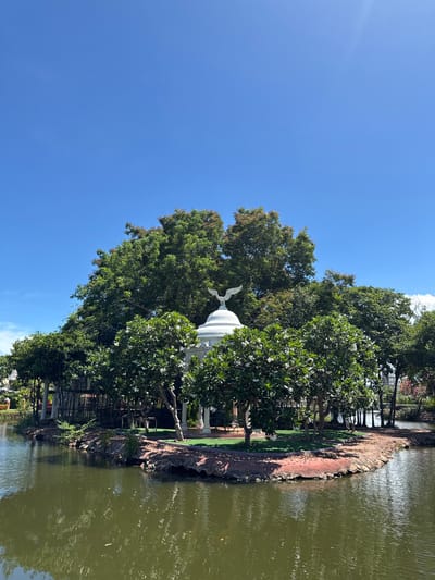 The image shows a partially obscured white pavilion with an eagle statue on top, set amidst trees and located on the edge of a body of water. The location is the Bang Pu Subdistrict Municipality, Thailand, evidenced by the typical Thai architecture style and local vegetation. A clear blue sky is overhead. The water shows some rippling and reflections, suggesting slight wind. Vegetation lines the water's edge, with trees of varying sizes including some with white blossoms. The pavilion's base is not fully visible due to the trees, but its general structure is evident. A small walkway or path runs along the water's edge in the vicinity of the pavilion. There are no people visible in the image.