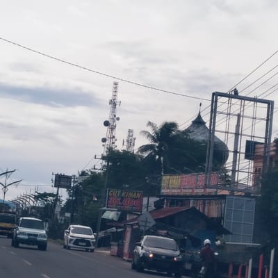 This image captures a bustling street scene in Sentosa, Indonesia, under an overcast sky. The foreground features a paved road with active vehicle traffic, including a white SUV, a yellow truck, a white sedan (with license plate BG 1962 AM), a black car, and a person on a motorcycle wearing a white helmet.
Along the right side of the road, various roadside structures are visible, appearing to be small shops or stalls. Prominent signage features the text "CUT KINAN 35.000" on a red and yellow sign, indicating a price, possibly for a service or product. Another larger sign, partially obscured, also shows "SEMUA 35.000" (meaning "All 35.000"), with smaller price denominations such as "10.000", "15.000", "25.000", and "35.000" below it. A partial word "SRRC" is also faintly visible on a structure.
In the mid-ground and background, several tall, metal communication towers equipped with multiple antennas stand against the cloudy sky, suggesting a significant telecommunications presence. Lush palm trees and other green foliage are scattered throughout the scene. Behind a large, empty metal billboard frame on the right, the distinct dome and finial of a building, characteristic of a mosque, can be observed. The overall impression is a typical day in a semi-urban or urban area of Indonesia, characterized by mixed commercial and residential elements, essential infrastructure, and daily activities.