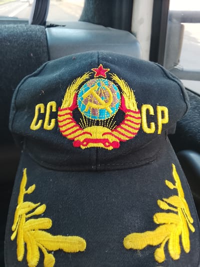 A black cap with a Soviet emblem embroidered on the front sits on a dark car seat. The emblem features a red star, a globe with a hammer and sickle, and yellow and red wheat stalks. Yellow lettering on the cap reads "CC CP." The cap is positioned on a dark fabric car seat, with the interior of the car visible in the background, including a window and what appears to be a road or highway. The image quality is adequate.