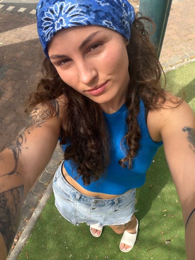 A young person, likely female, is captured in a high-angle, low-perspective selfie, seemingly reclining or lying down outdoors in Kiryat Motzkin, Israel. She looks directly at the camera with a relaxed expression and a slight smile.
She has long, dark, curly hair that flows around her head and shoulders. Her attire includes a blue bandana with a white floral or paisley pattern, a ribbed blue tank top, and light-wash, distressed denim shorts. She wears white slide sandals on her feet. A silver nose ring is visible, and multiple tattoos are prominent on her arms, shoulders, and potentially her lower back or side.
The background shows a paved or brick pathway on the left and artificial green turf or grass on the right. A dark green metal rail or bench structure runs horizontally in the upper left. The scene is brightly lit by natural sunlight, indicating it is daytime, with shadows cast clearly on the ground. There is no visible text in the image.