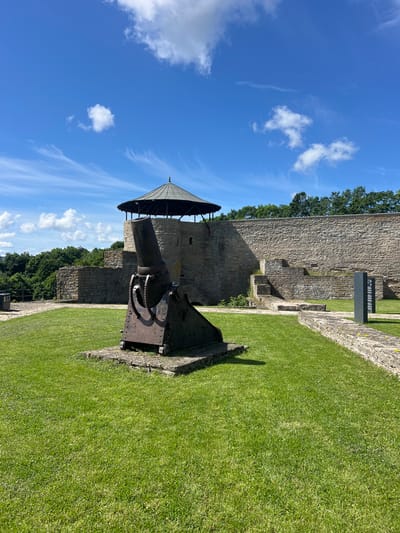 This image, taken in Narva, Estonia, depicts a historical outdoor scene on a sunny day. The primary subjects are a weathered, dark-colored cannon situated on a grassy area, and the stone walls of what appears to be a fortification or castle. The cannon is positioned on a metal base with visible gears and is pointed towards the left of the frame. Above and behind the cannon, a conical, dark-colored roof structure is visible, supported by wooden beams.
The background is dominated by the ancient, rough-hewn stone walls of a building, suggesting a historical site. Trees with lush green foliage are visible beyond the walls, and a bright blue sky with scattered white clouds fills the upper portion of the image. The grass in the foreground is vibrantly green and well-maintained, casting shadows from the cannon and the walls.
The overall atmosphere is tranquil and evokes a sense of historical significance. The time of day appears to be mid-morning or early afternoon, indicated by the clear sky and the angle of the shadows. No people are visible in the frame. There is no discernible text in the image.