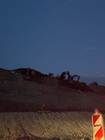 The image shows a construction site at dusk in what is purportedly Prijeradi, Montenegro. Earth-moving equipment, including excavators, is visible on a dirt hill. A white truck is parked at the base of the hill. The sky is dark blue. A construction barrier marked "BEMAX" is positioned on the right side of the image. The ground near the barrier is covered in gravel.