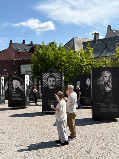 An outdoor photography exhibition is taking place on a cobblestone plaza in the city of Lund, Sweden. The exhibition features large, black-and-white portraits displayed on tall, freestanding panels. The portraits are of various individuals, including a woman wearing a hat and the singer Zara Larsson giving the middle finger. Several people are viewing the exhibition. In the foreground, a person with short hair and wearing a white shirt and striped pants is looking at a portrait and holding a white cup. To their right, a man wearing a striped shirt and brown pants is standing and looking at the same portrait, holding a small object in his hand. In the background, buildings with red brick and dark roofs are visible, along with lush green trees. The sky is blue with some white clouds. The overall atmosphere is casual and public, suggesting a cultural event or art display.