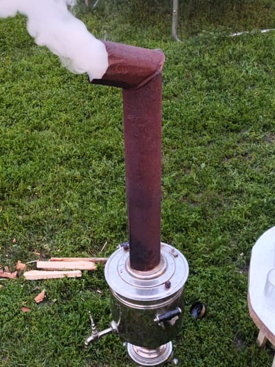The image shows a samovar with a rusted chimney emitting white smoke, set on a green grassy lawn. Several pieces of firewood are scattered on the grass near the samovar. To the right, a portion of a white table with a glass is visible. The setting appears to be outdoors, with hints of a rural or suburban environment. There are no people visible in the image. The overall scene suggests a moment of preparing or using the samovar for tea or hot water. The location of Новоурсаево, Russia, is not directly depicted or inferable from the visual elements within the image itself.