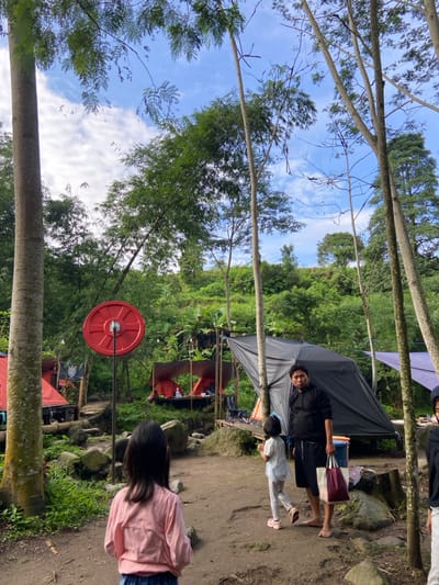 A person with a child stands outside a black tent near a path. Another person, wearing a pink shirt, has their back to the camera. A red weight hangs from a metal bar between the trees. The background consists of a dense green forest and a blue sky with white clouds visible through the treetops. Tall, slender trees line the perimeter.