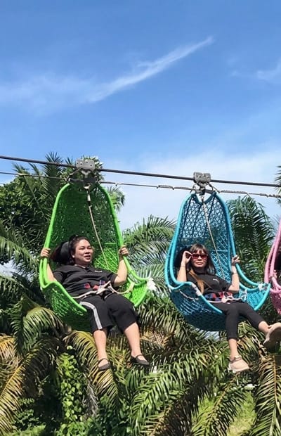 Two women are seated in colorful hanging swings suspended from a wire, against a backdrop of lush green palm trees and a clear blue sky with a wispy cloud. The woman on the left is in a green swing, wearing a black t-shirt and black pants. She is smiling and holding onto the sides of the swing. The woman on the right is in a blue swing, wearing a black t-shirt and black pants, and sunglasses. She is also holding onto the sides of her swing, with a relaxed posture. The swings appear to be part of an attraction, possibly a zipline or a scenic viewpoint, located in Bintang Meriah, Indonesia. The overall scene depicts a recreational activity in a tropical environment.