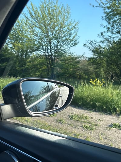The image shows a view from inside a car. The side mirror is prominently visible, reflecting another car. Outside the car, there are trees and vegetation under a blue sky. The setting appears to be a rural or semi-rural area, with a road visible alongside the vegetation. There is no explicit indication of the location being Kozar Belene, Bulgaria, in the image.