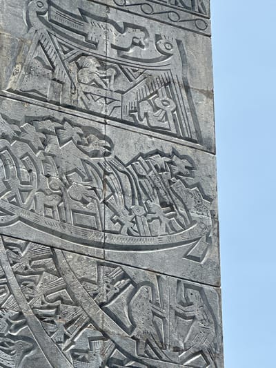 This is a close-up, low-angle, eye-level shot of a section of a tall, grey stone monument with bas-relief carvings. The monument is positioned in Ben Cau, Vietnam, against a clear, bright blue sky. The carvings depict stylized figures and scenes, appearing to be historical or mythical in nature. The stone has a rough texture and visible seams where different sections are joined. The carvings are intricate, with sharp lines and varying depths, creating shadows that emphasize the three-dimensional quality of the artwork. The overall impression is one of ancient artistry and monumental scale.