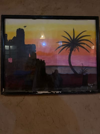 This is a framed painting of a desert sunset scene, likely depicting a location in Agafay, Morocco. The painting features a silhouette of ancient ruins on the left, with a bird in flight against a gradient sky of yellow, orange, and pink. A large palm tree dominates the right side of the composition, with its fronds spread wide. In the foreground, silhouetted figures are visible, suggesting human presence. A smaller palm tree is also depicted on the right. The painting is signed in the lower right corner. The artwork is displayed on a textured, earthy-toned wall.