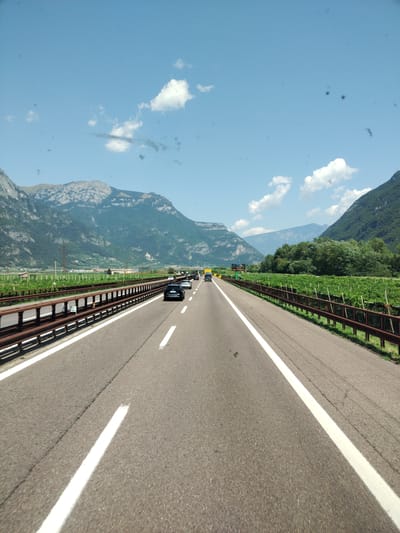 The image is a roadside view of a highway in Avio, Italy, on a sunny day with a clear blue sky dotted with white clouds. The highway, with asphalt paving and white lane markings, runs through a valley flanked by lush green mountains. A rusty metal guardrail separates the highway from a vineyard on the right side of the road. Several cars are visible on the highway, driving in the same direction as the viewer. The scene is devoid of any people, and the overall impression is one of travel and natural beauty.